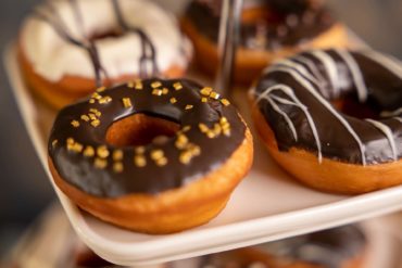 Chocolate glazed donut with gold sprinkles on a cake stand, more glazed donuts behind