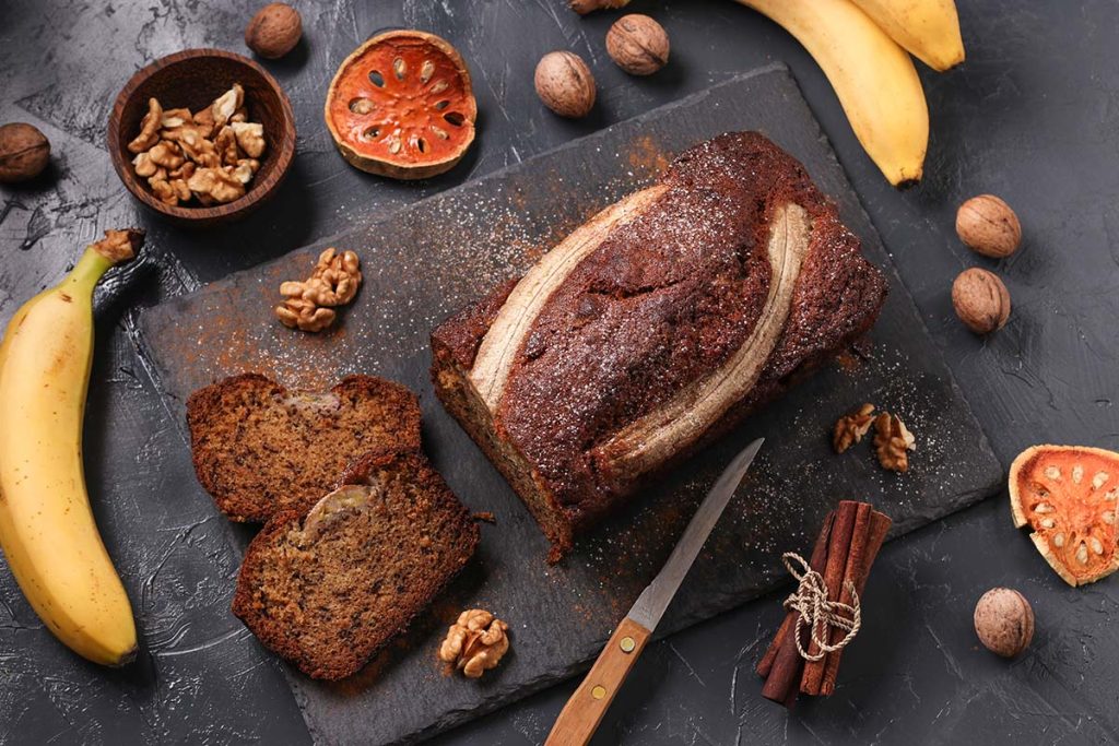 A beautifully presented banana bread loaf on a rustic slate board, sliced to reveal its moist texture. The bread is garnished with a topping of banana slices, surrounded by walnuts and whole bananas. The setting includes warm, earthy tones with scattered walnuts and cinnamon sticks, creating a cozy and inviting atmosphere perfect for showcasing a homemade banana bread recipe.