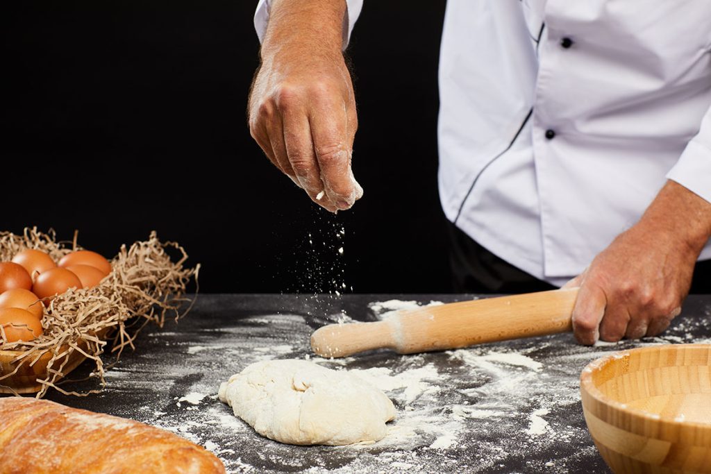A baker sprinkles flour onto a dough ball while preparing to roll it out with a rolling pin. In the background, there are fresh eggs in a basket and a loaf of bread on a floured countertop.