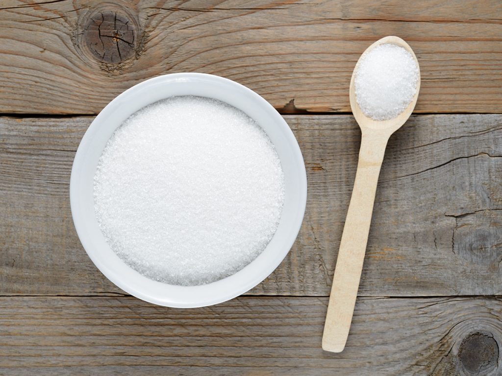 A bowl filled with fine granulated sugar next to a wooden spoon also containing sugar, all set on a rustic wooden table. The sugar appears white and crystalline, ready for use in baking or cooking.