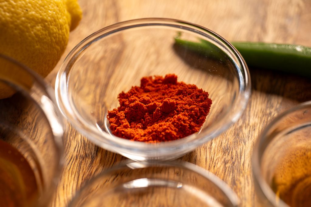 Close-up image of a small glass bowl filled with sweet paprika powder on a wooden surface, accompanied by a lemon and a green chili in the background. The vibrant red color of the paprika contrasts with the natural textures and colors of the other ingredients.