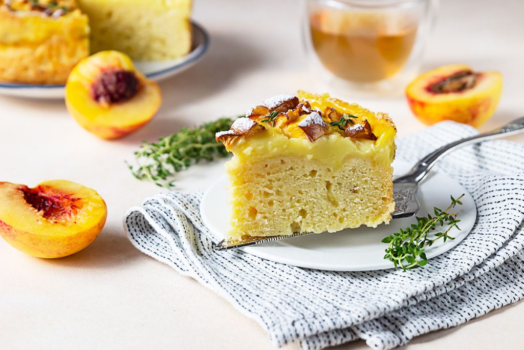 A slice of fluffy cake with a golden crust, topped with powdered sugar and garnished with fresh thyme. The cake is served on a white plate with a fork, set on a striped cloth. In the background, there are fresh peach halves, a glass of tea, and more cake on a plate.