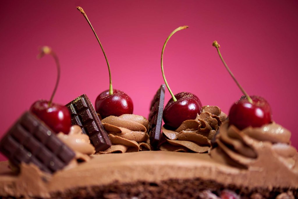 The image features a close-up of a decadent chocolate cake topped with swirls of rich chocolate frosting, juicy red cherries, and pieces of dark chocolate. The vibrant red background enhances the indulgent and luxurious feel of the dessert, making it an irresistible treat that is perfect for special occasions or celebrations.