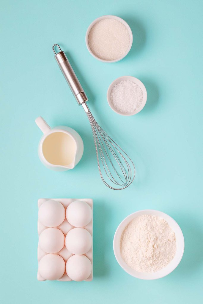 The image shows a flat lay of baking ingredients and tools on a light turquoise background. The items include a whisk, a carton of six eggs, a small pitcher of cream, a bowl of flour, a bowl of sugar, and a bowl of salt. The composition is clean and minimalist, highlighting the essential ingredients typically used in baking.