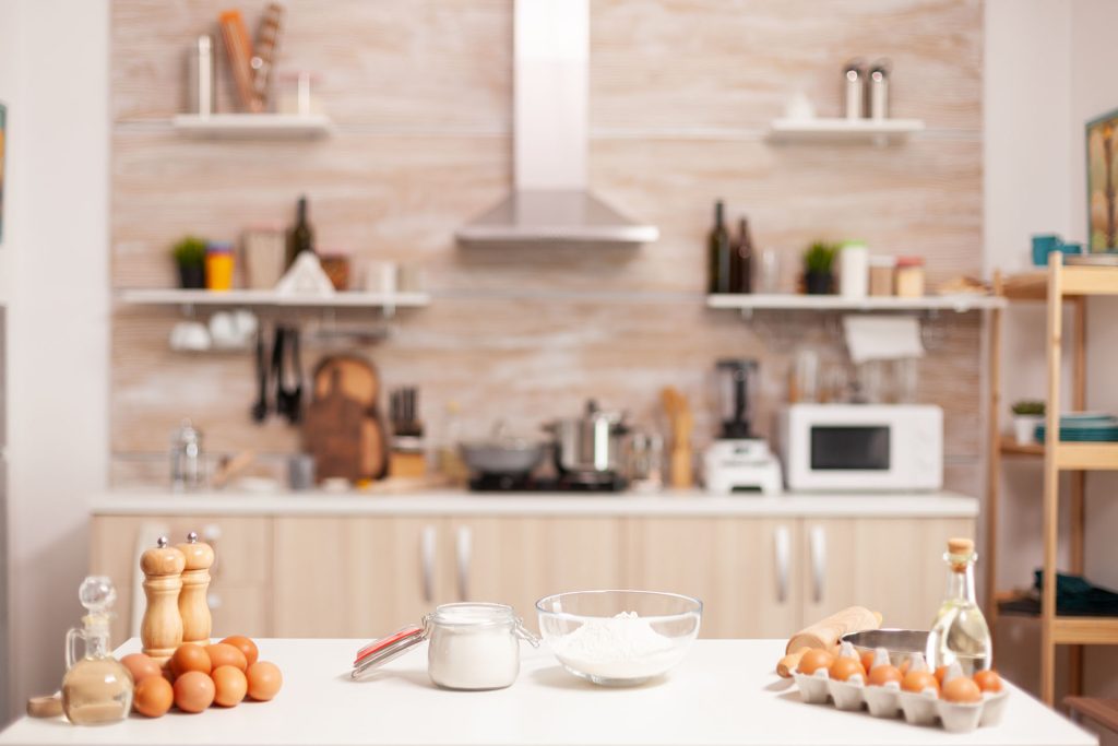 This image showcases a bright and modern kitchen with a focus on a clean, white countertop in the foreground, which is neatly arranged with baking ingredients. On the countertop, there are several items, including a carton of eggs, a glass jar of flour, a small bottle of oil, and a bowl of sugar. Additionally, there are wooden salt and pepper mills, a rolling pin, and a whisk, all set up, suggesting preparations for baking. The kitchen in the background features wooden cabinets, open shelves with various kitchen utensils, a microwave, and a stove, all contributing to the cozy, organized atmosphere of a home kitchen ready for cooking or baking.