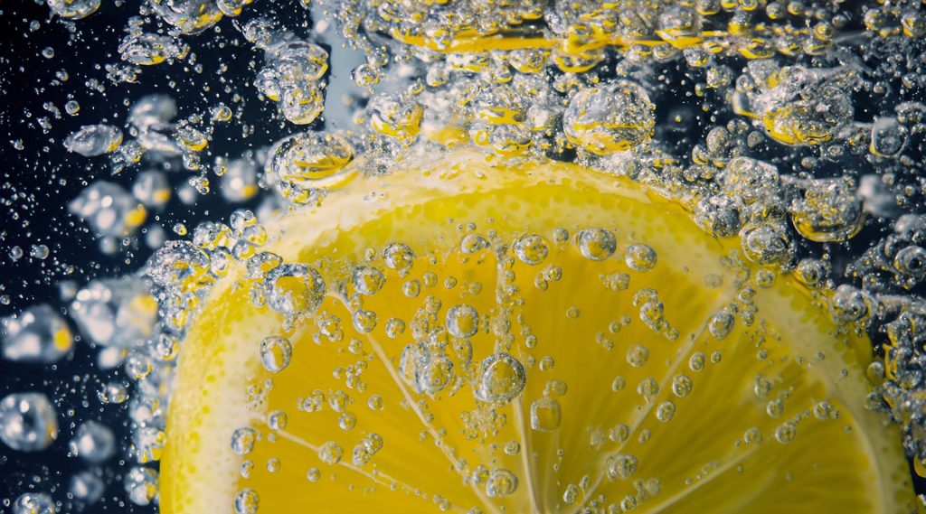 Close-up of a vibrant yellow lemon slice submerged in sparkling water, surrounded by effervescent bubbles. The image captures the refreshing and zesty essence of lemon-infused carbonated drinks, emphasizing the freshness and fizz. The high-definition macro shot highlights the intricate details of the bubbles and the texture of the lemon, making it an ideal visual representation for discussing the benefits of homemade sparkling water or enhancing beverages with citrus flavors.