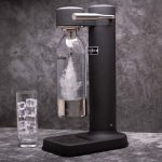 Close-up of a sleek, modern black soda maker with a carbonating bottle in place, producing sparkling water. The appliance is placed on a dark, textured surface with a glass filled with ice cubes beside it. The minimalist design and premium build quality are highlighted, ideal for home use. The image emphasizes the ease of creating sparkling water at home, making it a perfect fit for any kitchen aesthetic.