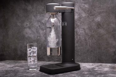 Close-up of a sleek, modern black soda maker with a carbonating bottle in place, producing sparkling water. The appliance is placed on a dark, textured surface with a glass filled with ice cubes beside it. The minimalist design and premium build quality are highlighted, ideal for home use. The image emphasizes the ease of creating sparkling water at home, making it a perfect fit for any kitchen aesthetic.