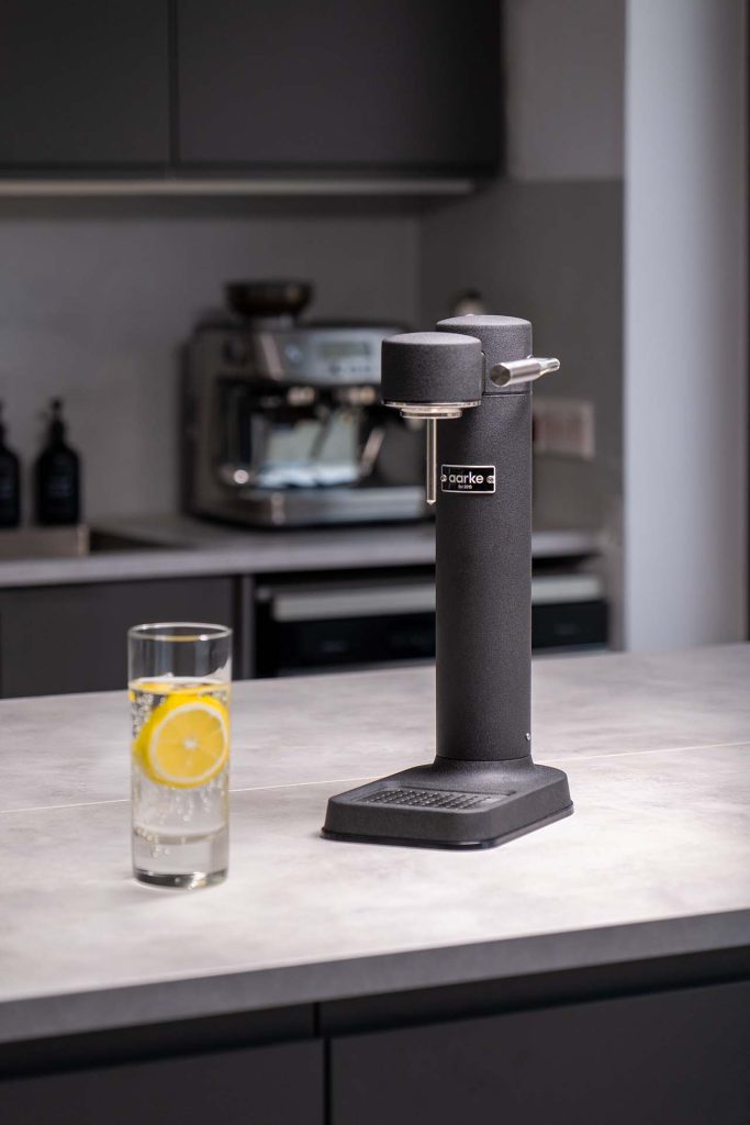 Modern kitchen setting featuring a sleek black Aarke soda maker prominently displayed on a light-colored countertop. A tall glass of sparkling water with a slice of lemon sits beside the appliance, ready to be enjoyed. The background includes a sophisticated coffee machine, adding to the contemporary and stylish atmosphere of the kitchen. This image emphasizes the soda maker's elegant design and its ability to seamlessly fit into any modern home decor.