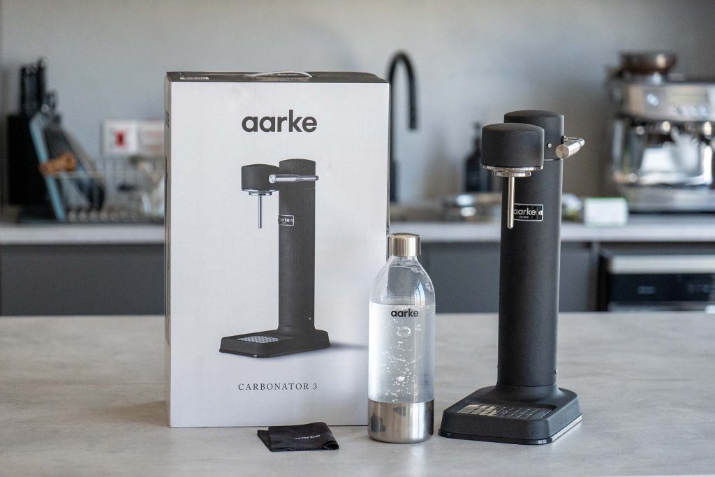 Image of an Aarke Carbonator 3 in a modern kitchen setting. The carbonator, in a sleek black finish, is placed on a countertop next to its packaging box. A transparent bottle filled with carbonated water is positioned in front of the carbonator, showcasing its use. The background features kitchen appliances and utensils, adding context to the product's functionality.