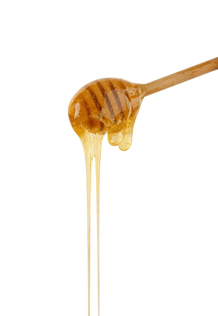 A wooden honey dipper drips golden honey against a white background, capturing the thick, smooth flow of the honey as it slowly streams down. This image perfectly illustrates the use of honey as a natural sweetener in baking, ideal for adding moisture and rich flavor to cakes, cookies, and bread