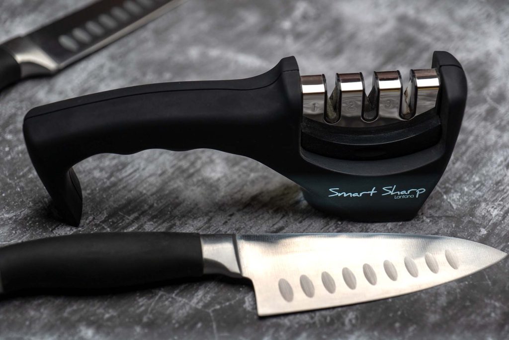 A close-up image of a black handheld knife sharpener labeled 'Smart Sharp' with three sharpening slots. A Japanese kitchen knife with a black handle is placed next to the sharpener on a textured surface, emphasizing the tools used for maintaining a sharp blade in the kitchen.