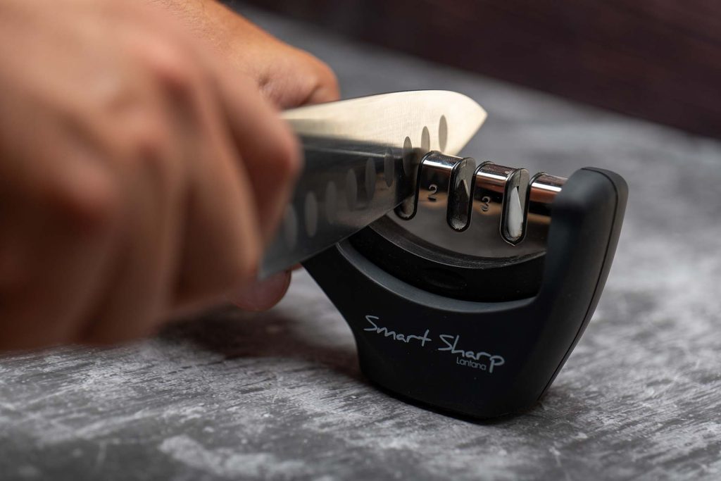 A close-up image showing a person using a 'Smart Sharp' handheld knife sharpener to sharpen a kitchen knife. The knife is being pulled through one of the sharpening slots, emphasizing the hands-on process of maintaining a sharp blade.