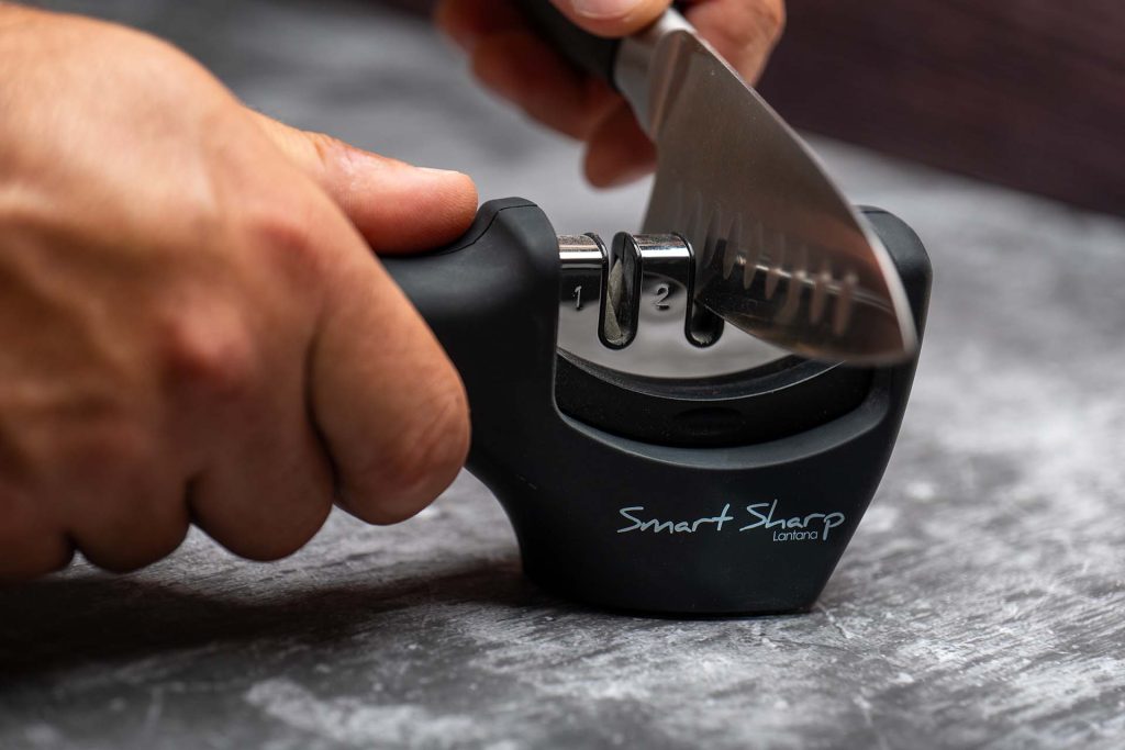 A close-up image of hands holding a 'Smart Sharp' knife sharpener while pulling a kitchen knife through one of the sharpening slots. The image focuses on the hands-on process of effectively sharpening a knife to maintain its edge.