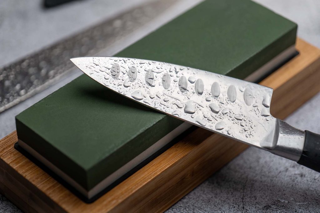 A detailed image showing a Japanese-style kitchen knife, covered in water droplets, resting on a green whetstone placed on a wooden base. The image highlights the process of knife sharpening and the high-quality blade.