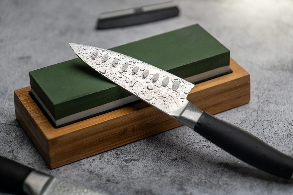 A close-up image of a Japanese-style kitchen knife with a hammered blade resting on a green whetstone, set on a wooden base, highlighting the sharpening process and the texture of the blade.