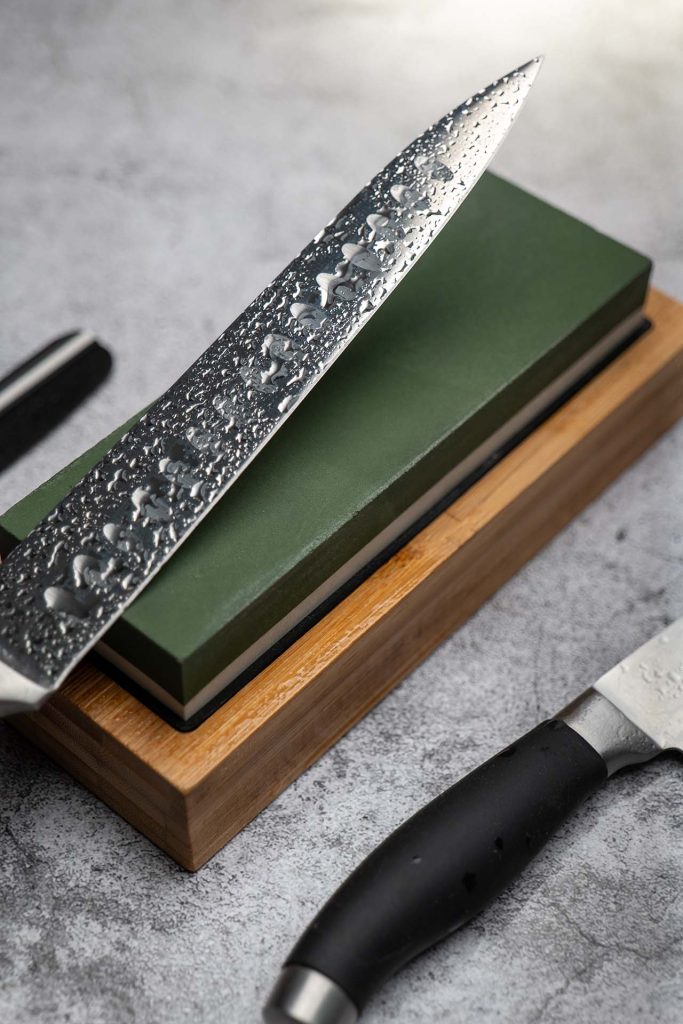 A close-up image of a long Japanese kitchen knife, featuring a hammered texture and water droplets, placed on a green whetstone on a wooden base. The knife's sharp edge is prominently displayed, emphasizing the sharpening process.