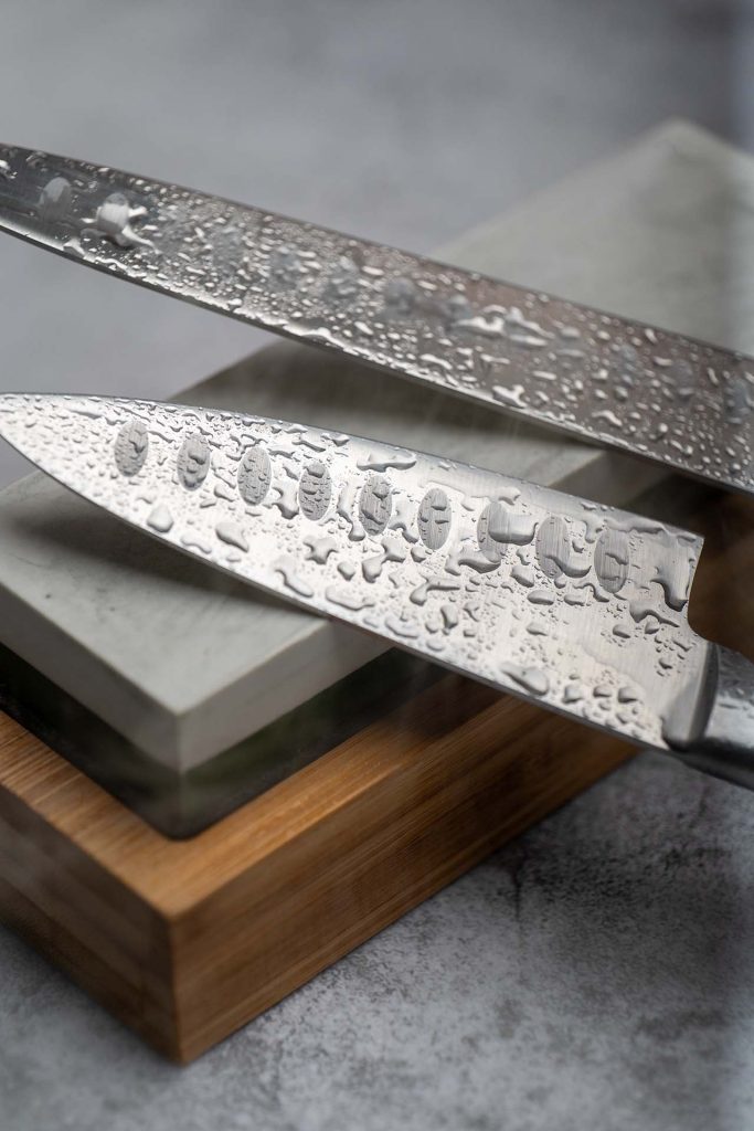 A close-up image of two Japanese kitchen knives, covered with water droplets, resting on a dual-sided whetstone. The image highlights the sharpening process and the high-quality craftsmanship of the knives.