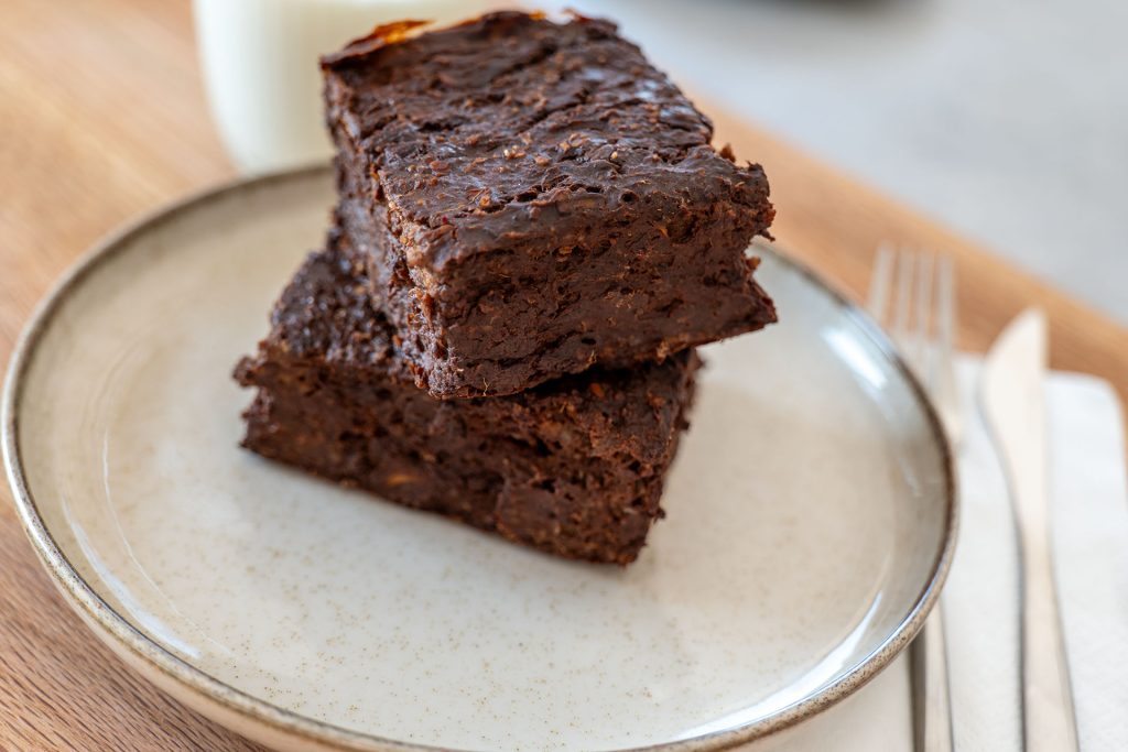 The image shows two square pieces of chocolate brownies stacked on a speckled ceramic plate. The brownies have a rich, dense texture, indicating a moist and hearty composition. In the background, a glass of milk is partially visible, and to the side, there are a fork and knife placed neatly on a napkin. The setting is casual, with a light wooden surface and natural lighting