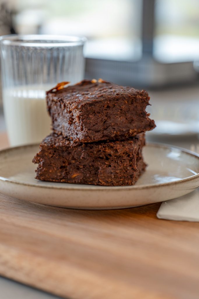 Close-up of two fudgy and rich sweet potato brownies stacked on a ceramic plate, paired with a refreshing glass of milk in the background. A perfect guilt-free dessert made with wholesome ingredients.